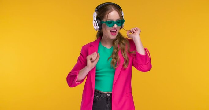 Trendy colorful dressed Woman is dancing in Headphones on yellow Background. Amazing Girl looking happy and satisfied while is listening to Music isolated on Studio. Concept: Musically Apps, Emotions.