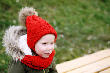 Cute little smiling girl in knitted woolwn cap and scarf sitting on a bench outdoors