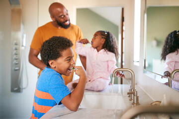 Father Helping Children To Brush Teeth In Bathroom At Home