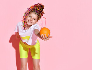 Smiling girl in bright yellow shorts and white t-shirt leaning forward proposing spectator cocktail in a fresh orange, isolated on pink