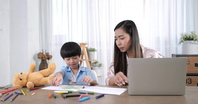 Happy asian mother works from home office with kid. woman using laptop. Cute child drawing in mother notebook. Freelancer workplace at living table. Female business, career. Lifestyle family mom