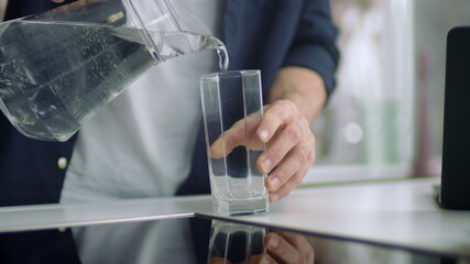 Closeup male hands pouring water into glass from jug at kitchen background.