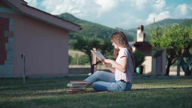 Smiling female student sitting on the grass near the house among the books and reading story. Concentrated girl reading book, turning pages and analyzing information. Preparation for exam outdoors