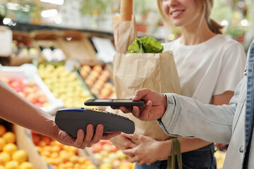 Close-up of young couple paying for fresh products with smartphone at organic food market
