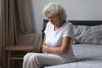 Unhealthy senior grey haired woman awakened woke up sit on bed in bedroom alone touch abdominal,...
