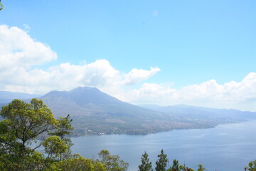 Bali, Indonesia. Batur volcano with caldera and Agung volcano, with water lake river, blue sky and clouds hanging and green vegetation trees.