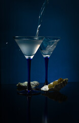 Clear liquid is pouring into martini glass with a splash,  and olives are lying in the oyster shell on a dark blue background.