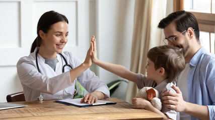 Happy young pediatrician in white uniform giving high five to small child boy sitting on fathers...