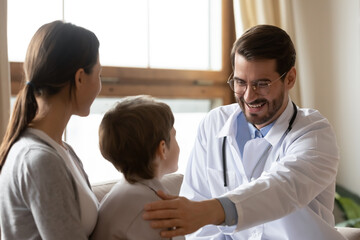 Fototapeta na wymiar Head shot happy friendly pediatrician in eyewear and white coat supporting little patient coming to meeting with mother. Emotional young doctor stroking shoulder of small kid boy at clinic visit.