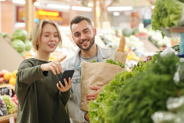 Positive young woman using smartphone app for purchasing and pointing at greens to boyfriend while...