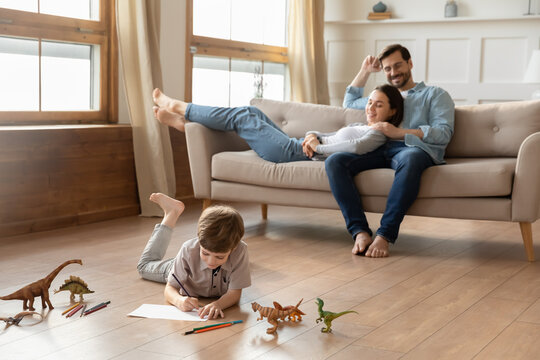 Smiling loving young bonding couple parents relaxing on comfortable sofa, watching little kid son drawing pictures in album, lying on wooden floor in living room, happy family weekend pastime. - Powered by Adobe