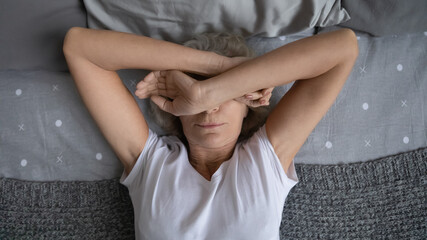 Top view aged woman lying on bed woke up at night due noisy neighbors. Mature female cover face with hands suffers from insomnia sleep disorder, has restless obsessive thoughts keep her awake concept - obrazy, fototapety, plakaty
