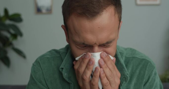 Close up view of ill young man blowing nose with wipe while sitting on couch at cosy room. Bearded guy having cold and using paper handkerchief. Concept of healthcare and illness