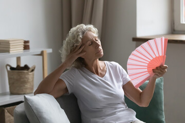 Overheated senior woman relax sit on sofa waving orange peach colour fan cool herself, feels unwell due unbearable hot weather, discomfort and hormonal changes, flat without air conditioner concept