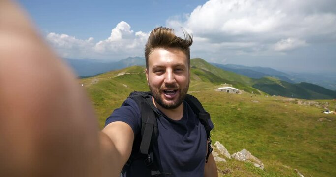 A carefree male tourist with backpack is making a selfie or technology video call to friends or relatives just reached a peak while hiking in the middle of hills surrounded by green nature.