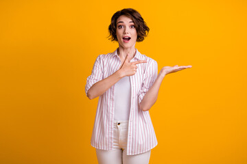 Photo of pretty young lady point direct finger hold palm empty space staring open mouth appartment...