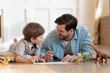 Happy small boy lying on floor with caring dad, enjoying funny conversation, discussing hand drawn pictures in living room. Emotional positive father involved in funny domestic activity with son.