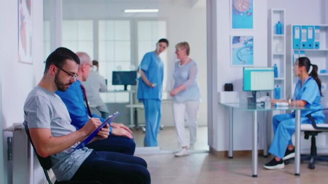 Man filling paperwork in hospital waiting area before doctor examination. Nurse inviting senior man in consultation room. Assistant in blue uniform working on computer at reception.