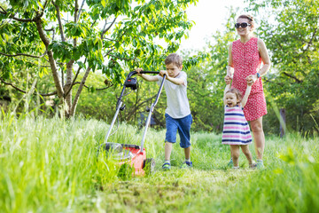 Young boy mowing grass with lawn mower, mother and sister encouraging him