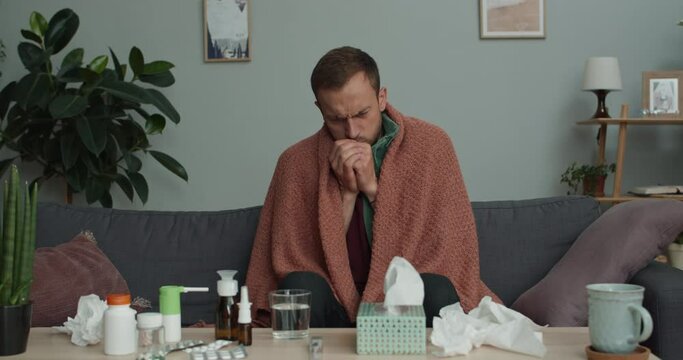 Bearded guy covered in blanket being ill while sitting on sofa at home. Man in 30s coughing loudly and s while getting sick and taking cure at home. Concept of healthcare and illness