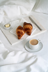 Breakfast with croissants on the napkin, knife and butter on the marble tray with a cup of coffee on the white bed with blanket. Cozy morning.