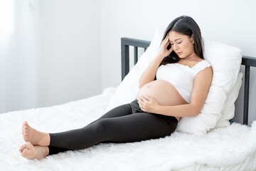 Beautiful Asian pregnant woman show action of stress and headache lie on bed with morning light....