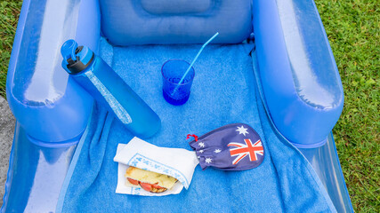 Composition in blue shades of a picnic on the grass with inflatable mattress, water bottle, sandwich, glass with straw and Australian flag
