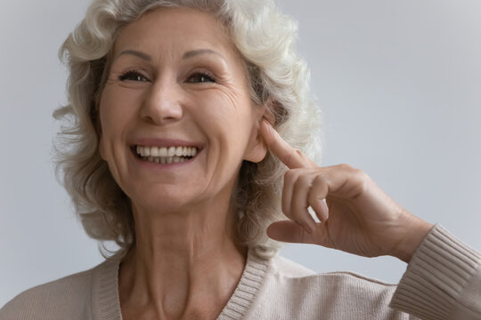 Close up image elderly woman point finger to ear feels happy by new modern comfort hearing aid device. Age related hearing loss, services of clinic ad for old patient diagnosis and treatment concept