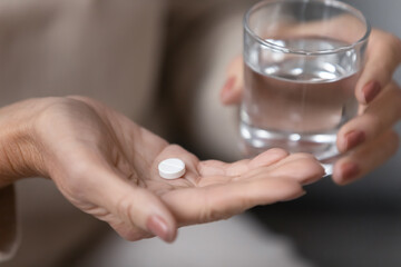 Senior woman hands holds round white pill and glass of still water close up view. Senile diseases prevention, cholesterol control tablet, cardiovascular heart disease treatment for old people concept