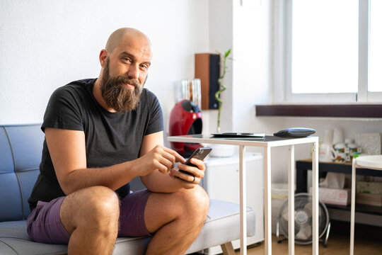 Hombre calvo con camiseta gris oscura, pantalón corto púrpura, y barba, mirando a cámara, sonriendo, con smartphone, sentado en un sillón en su oficina, con luz de día que entra por la ventana