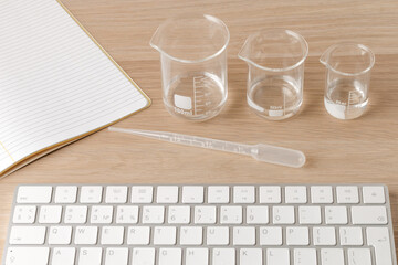 glass measuring beakers standing gradually on table and plastic pipette in front of a white computer keyboard, a segment of a ruled notebook laying, idea of taking precise measurements, research