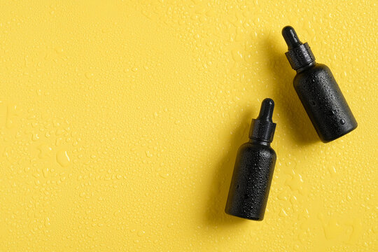 Wet black cosmetic dropper bottles in droplets of water on yellow background. Luxury essential oil packaging template, beauty product for hair or body care.