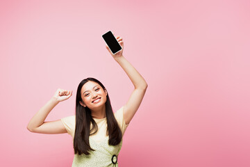 smiling asian girl with clenched fist holding smartphone with blank screen isolated on pink