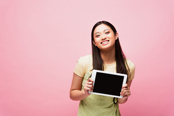 Fototapeta premium cheerful asian girl holding digital tablet with blank screen isolated on pink