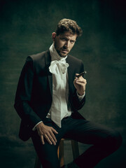 Brutal smokes cigar. Young man in suit as Dorian Gray isolated on dark green background. Retro...