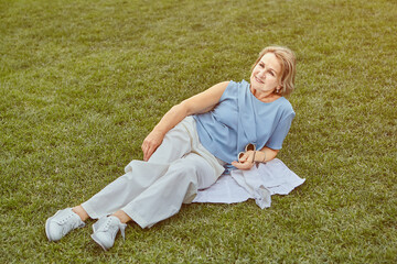 Aged cheerful woman is lying on grass in park.
