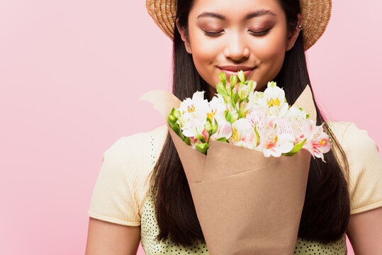positive asian girl in straw hat smelling flowers isolated on pink