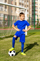 a small football player with a ball stands on a green football field in the goal