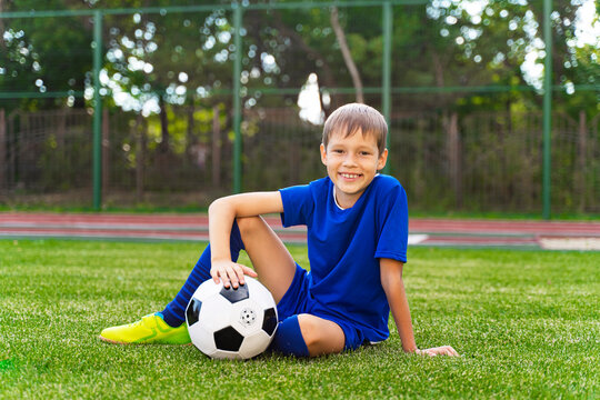 A little boy sits on a green soccer field, a soccer ball