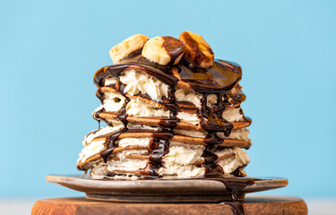 Stack of pancakes with cream, chocolate and bananas in a plate on a light blue background....