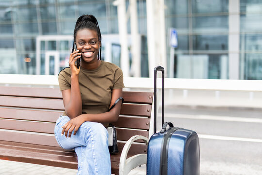 Portrait Of Happy Young African American Woman Sitting With Suitcase And Talking With Mobile Phone At The Airport