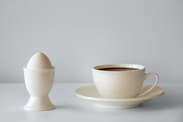 Boiled egg in an egg Cup and tea in a white Cup. White dishes on a white background. Breakfast consists of a boiled egg and a Cup of tea.
