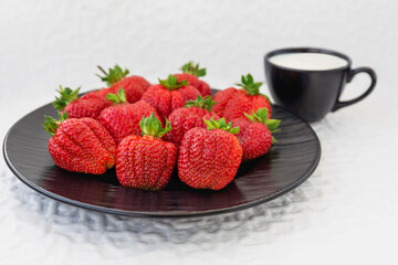 Strawberries on a black plate and a Cup of milk. A black dish with a large ripe fresh strawberry on the table. Selective focus. White background