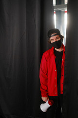 Emotional young man 25-30 years old in a black protective mask, a cap and a red jacket holding a megaphone against a dark background.