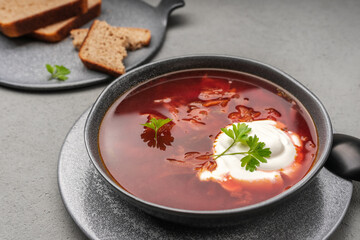Traditional borscht with sour cream and parsley in a grey tureen next to slices of bread and wholemeal flour on a grey concrete surface