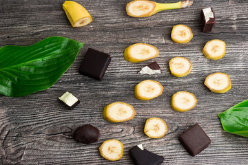 Slices of organic yellow bananas, chocolate candies and green leaves on wooden background - 369314244