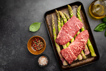 Raw Steak with green asparagus on tray on black background, top view