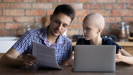 Focused young Caucasian couple with sick cancer patient woman work on laptop paying bills online,...