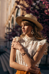 Elegant woman wearing straw hat, big earrings, golden wrist watch, lace blouse posing in street. Outdoor fashion portrait