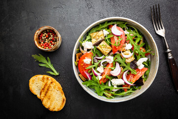 Eggplant salad with tomatoes, arugula and feta cheese in a bowl on black background, top view